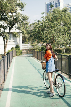 在城市公园的自行车道上，一个穿着橙色t恤和牛仔裤的长发女孩骑着自行车走着。