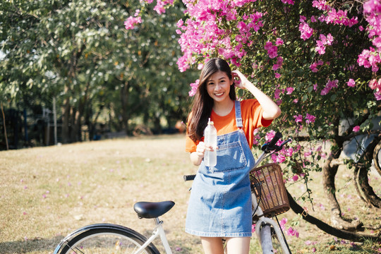 穿着橙色t恤和牛仔裤的长发女孩站在粉色的布干维尔旁，微笑着骑着自行车。