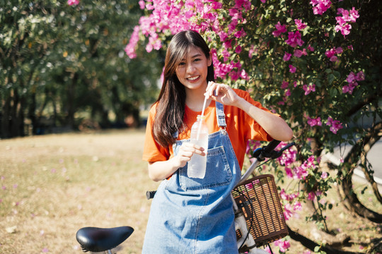 穿着橙色t恤和牛仔裤的长发女孩站在粉色的布干维尔边，手里拿着饮用水。