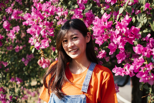 身着橙色t恤和牛仔裤套头衫的长发女孩在阳光下微笑着站在粉红色的布干维尔旁的肖像照。