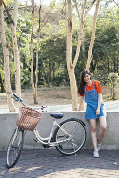 穿着橙色t恤和牛仔裤的长发女孩穿着套头衫，骑着自行车靠在鹅卵石路障上。