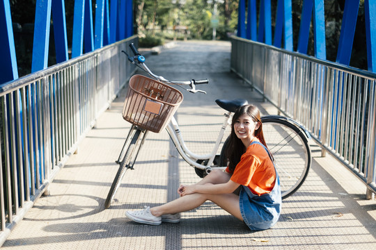 穿着橙色t恤和牛仔裤的长发女孩骑着自行车坐在蓝色的桥上。