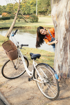 长发女孩穿着橙色t恤和牛仔裤套头衫躲在树后，在公园里把头伸出来。