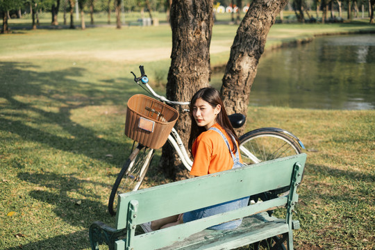 长发女孩穿着橙色t恤和牛仔裤套头衫坐在绿色公园的绿色长凳上。