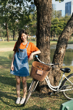 穿着橙色t恤和牛仔裤的长发女孩站在池塘边的树旁，骑着自行车。