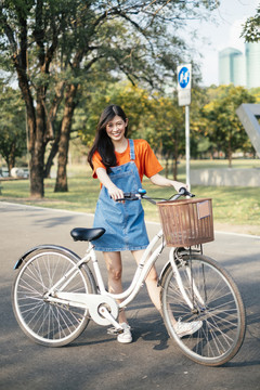长发女孩穿着橙色t恤和牛仔裤，站在公园的马路上骑着自行车。