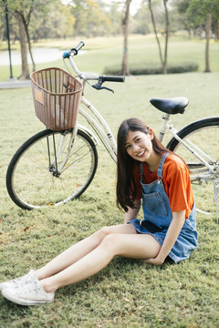 长发女孩穿着橙色t恤和牛仔裤，骑着自行车坐在公园的草地上。