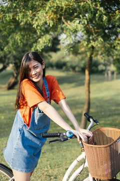 长发女孩穿着橘色t恤和牛仔裤，站在公园的田野里，与自行车垂直。
