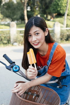 身着橙色t恤和牛仔裤套头衫的长发女孩在自行车上弯腰时展示橙色冰淇淋。