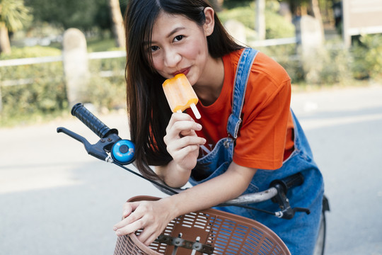 穿着橙色t恤和牛仔裤的长发女孩在自行车上吃橙色冰淇淋。
