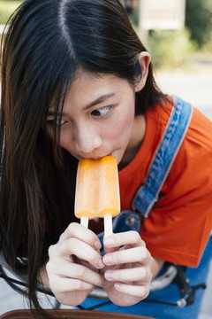 长发女孩穿着橙色t恤和牛仔裤在自行车上吃橙色冰淇淋，特写。
