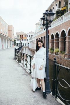 美丽的白衣长发女孩站在浪漫小镇河边的灯柱旁。