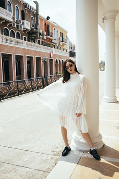美丽的长发女孩，穿着白色的裙子，在阳光下靠在白色的大柱子上。