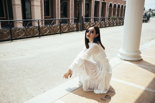 美丽的长发白裙女孩坐在阳光下的建筑在经典小镇。
