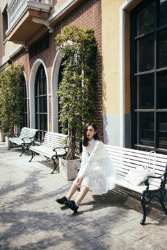 一个穿着白裙子的漂亮长发女孩坐在餐厅前的长凳上，和一棵树在一起。