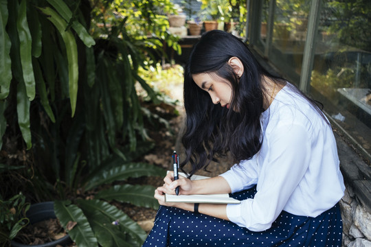 亚洲女性在笔记本上写字，用笔在窗边。在户外工作的自由记者。
