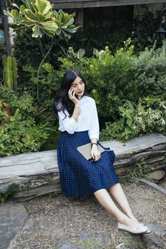 一位穿着白色连衣裙的亚洲年轻漂亮女子坐在原木上，讲着电话，手里拿着一本书。