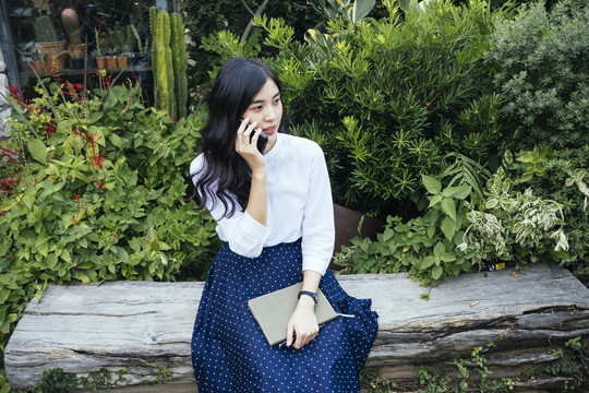 一位穿着白色连衣裙的亚洲年轻漂亮女子坐在原木上，讲着电话，手里拿着一本书。