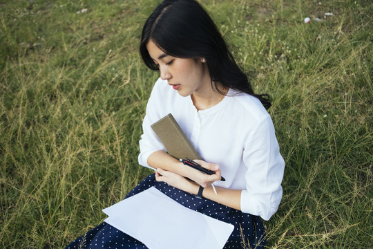 亚洲爱书的女人抱着一本书，坐在草地上。