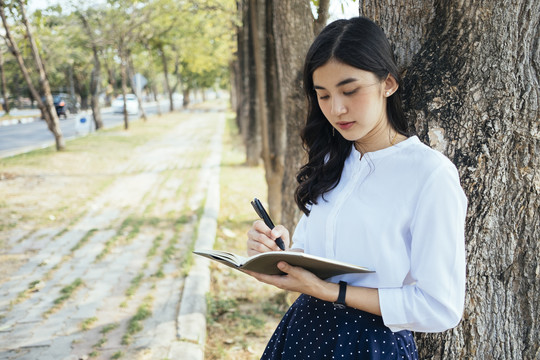 一位亚洲妇女靠在树上写着一本书。小说作家把这个故事写在一本书上。