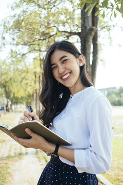 快乐的亚洲女人穿着白衬衫写着一本书，带着快乐的感觉，笑着笑着。小说作家微笑着写作的女孩。