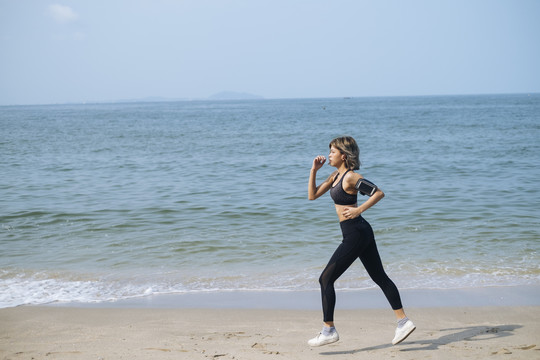穿着运动服的短发女人在沙滩上跑步，在阳光明媚的日子里穿着运动内衣和袖标，里面戴着智能手机。