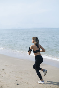 穿着运动服的短发女人在沙滩上跑步，在阳光明媚的日子里穿着运动内衣和袖标，里面戴着智能手机。
