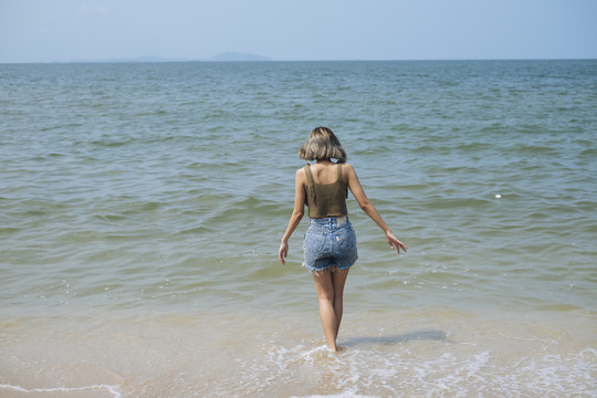 短发女孩穿着短牛仔裤喜欢在沙滩上散步，喜欢大海和海浪，赤脚。