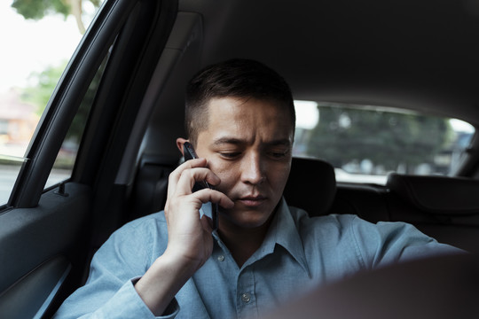 穿着蓝色衬衫的商人坐在汽车后座，在旅行中用手机看手表。