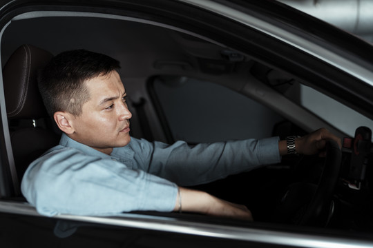 穿着蓝色衬衫的商人坐在车里的司机座位上，一只手放在车窗上，一只手放在方向盘上。