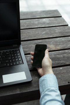 一张裁剪过的照片，是一名男子在木桌上的黑色笔记本电脑旁拿起智能手机的手。