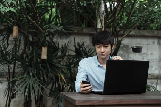 穿着蓝色衬衫的黑发男子坐在花园里，看着手机查看顾客的订单。