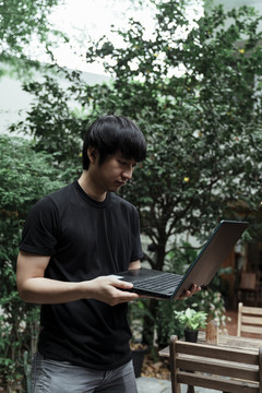 一个穿着黑色t恤的男人站在咖啡馆的花园里，手里拿着电脑。