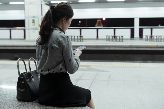 复制空间-亚泰女子坐在长凳上用智能手机听音乐，在火车站台上用耳机等火车回家。