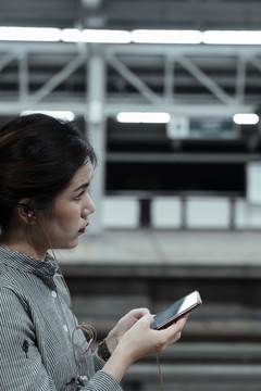 亚泰妇女站在火车站台上用智能手机听音乐侧视图。