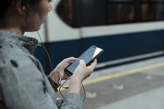 亚泰妇女站在火车站台上用智能手机听音乐侧视图。
