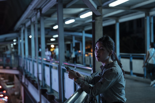 亚泰女子晚上用智能手机看着马路，站在天桥上。