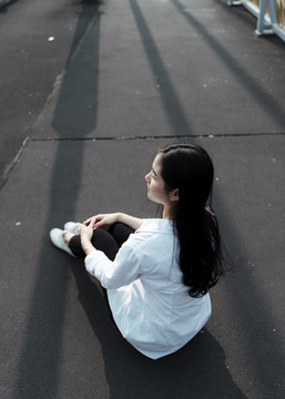 黑色长发女子穿着白衬衫坐在阳光下的黑色地板上。