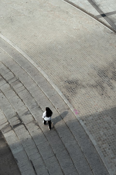 俯视图：身穿白衬衫的黑色长发女子走在广场的鹅卵石地板上。