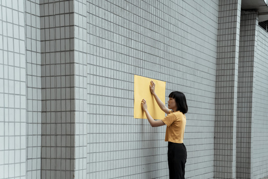 年轻的泰国亚裔女子深色短发，穿着橙色衬衫，在墙上贴上墙纸。户外。