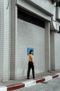 年轻的泰国亚裔女子深色短发身穿橙色衬衫站在街上，头后贴着蓝色墙纸。