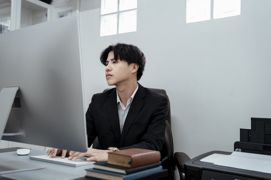 穿着黑色西装的亚洲商人坐在办公室的椅子上用电脑工作。