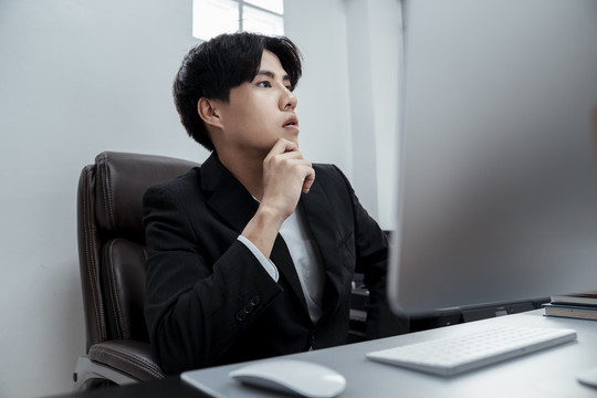 体贴的亚洲商人穿着黑色西装坐在电脑前的办公室椅子上工作。