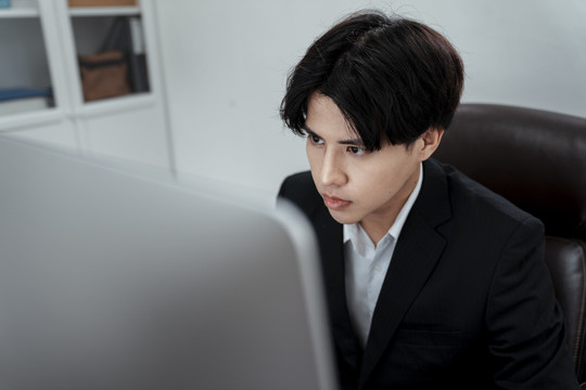 严肃的亚洲商人穿着黑色西装专注于看电脑。在办公室认真工作。