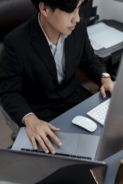 俯视图-身穿黑色西装的亚洲商人在办公桌上用笔记本电脑工作。