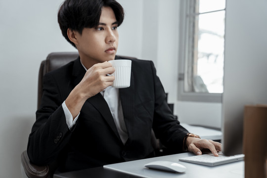 年轻英俊的亚泰商人身穿黑色西装，端着一杯咖啡在办公室里用电脑工作。