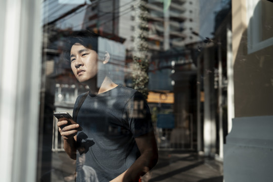 亚洲游客站在透明玻璃墙后用智能手机看街。