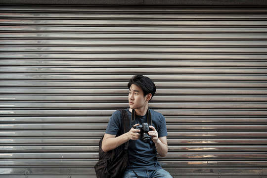 年轻英俊的亚洲摄影师坐在城市的钢卷帘门前-复印空间。