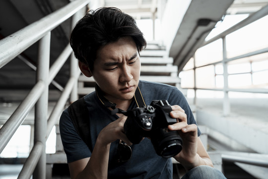 严肃的年轻英俊的亚洲摄影师坐在楼梯上看着相机里的照片，表情很不高兴。