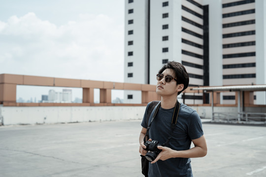年轻英俊的亚洲摄影师戴着太阳镜拿着相机走在屋顶寻找灵感。